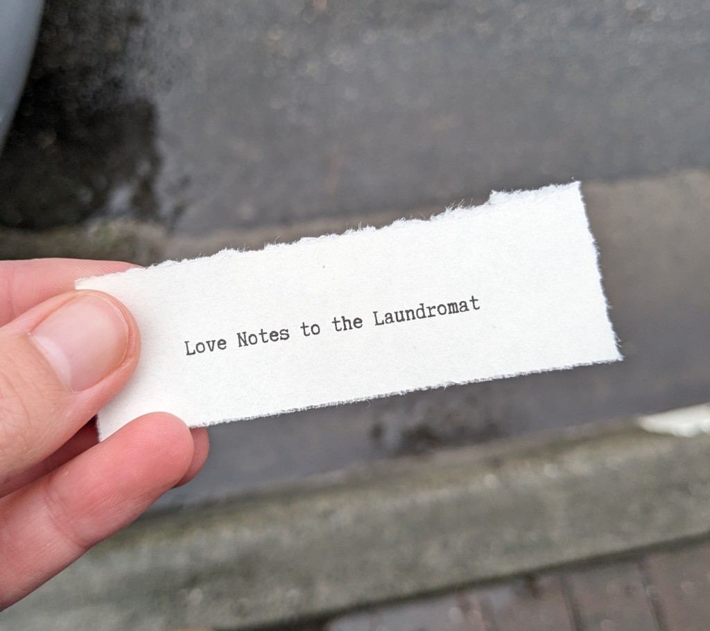 Hand holding a note that reads Love Notes to the Laundromat in a typewriter style font. 